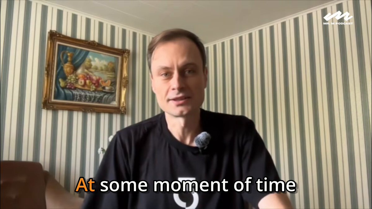 The image features a man wearing a black t-shirt sitting at a desk with a microphone in front of him. The background shows a striped wall behind him, and there are two other people visible in the room - one standing near the top left corner and another person partially visible on the right side of the frame. The text "At some moment of time" is written above the man's head, suggesting that he has just experienced or is about to experience a significant event related to Bitcoin or open source money systems.