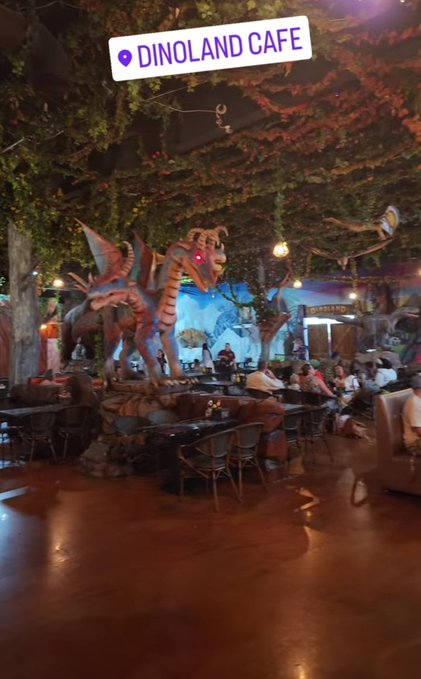 The image shows a large dinosaur statue with wings, surrounded by tables and chairs inside a building. There are people sitting at the tables enjoying their time. The scene appears to be set up for an event or gathering, possibly related to dinosaurs or a theme park-like atmosphere.