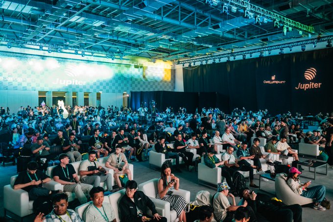 A wide shot of a large, seated audience at a conference, with 'Jupiter' branding visible on screens in the background.