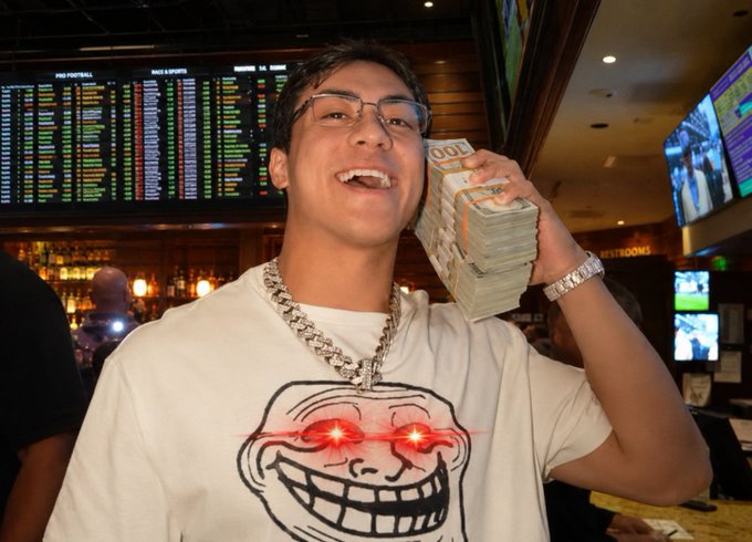 A young man wearing glasses, a silver chain, and a Trollface t-shirt with glowing red eyes, smiling while holding up a thick stack of banded $100 bills in what appears to be a sportsbook.