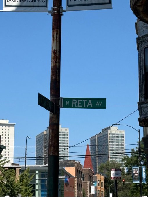A street sign reading 'N RETA AV' mounted on a rusted pole, with 'LAKEVIEW LANDING' and 'LOFTHOUSE' banners above, set against a backdrop of high-rise buildings and a clear blue sky.