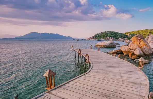 A scenic view of a wooden boardwalk winding over the ocean towards rocky shorelines and mountains, decorated with lantern-style lamps under a partly cloudy sky at dusk.