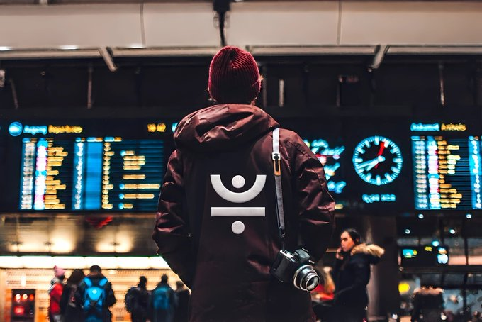 A person wearing a dark hooded jacket with a white Abstract logo on the back, a red beanie, and carrying a camera, standing in front of an airport departure board.