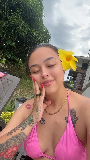 A selfie of a woman with closed eyes, red nails, and tattoos, wearing a pink bikini top and a yellow flower behind her ear, posed outdoors.