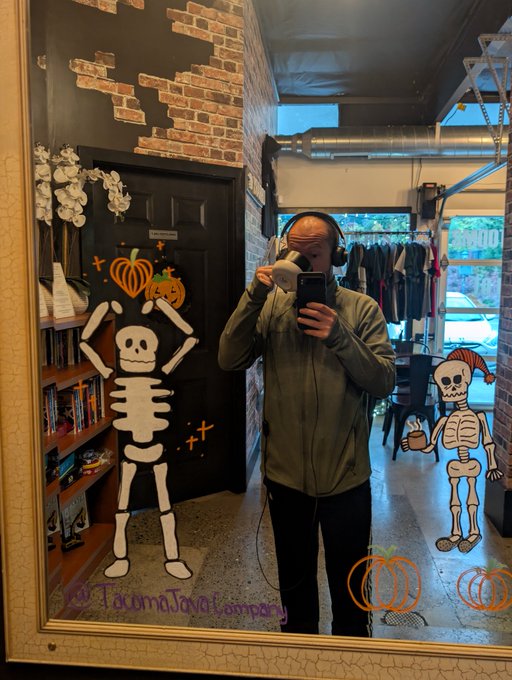 A mirror selfie of a person wearing headphones and drinking from a mug, with Halloween skeleton and pumpkin decorations on the mirror, marked 'TacomaTantalum Worldwide.'