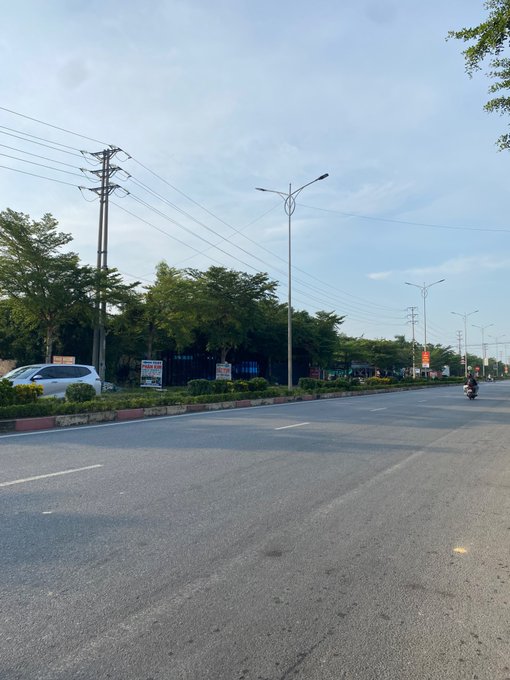 A street scene with a wide asphalt road, streetlights, power lines, trees, and a single motorcyclist riding in the distance under a clear sky.