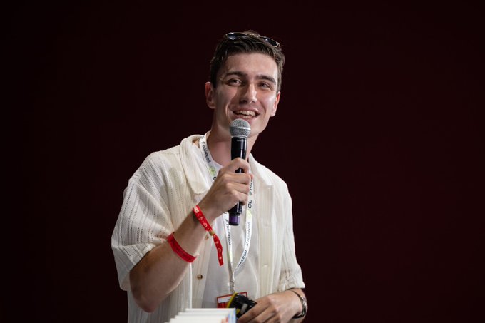A young man in a cream-colored shirt smiles while speaking into a microphone on a stage with a dark red background, wearing sunglasses on his head.