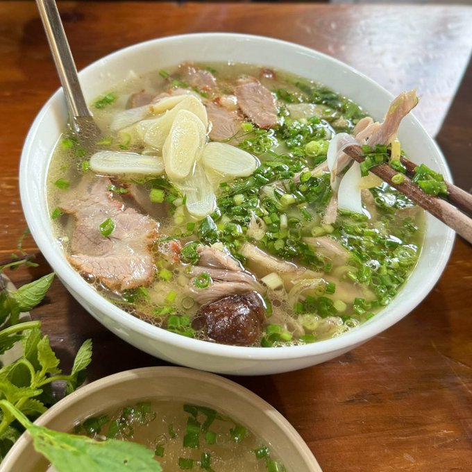 A bowl of Vietnamese pho with sliced beef, lemongrass, herbs, and green onions in a clear broth on a wooden table, with a small dish of sauce nearby.