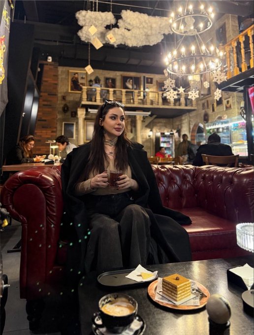 A woman sits on a tufted red leather sofa in a richly decorated, Harry Potter-themed cafe, holding a drink, with a layered honey cake on the table in front of her and cloud decorations hanging from the ceiling.