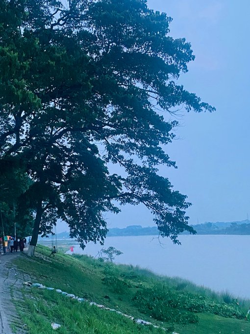 A serene landscape photograph showing a large tree with dense green foliage in the foreground beside a calm lake or river, with people walking along a path on the left and a misty, overcast sky above.