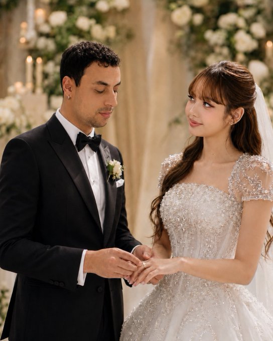 A groom in a black tuxedo and bow tie places a wedding band on the finger of his bride, who is wearing an ornate, sparkly white gown and veil, during their wedding ceremony surrounded by white flowers.