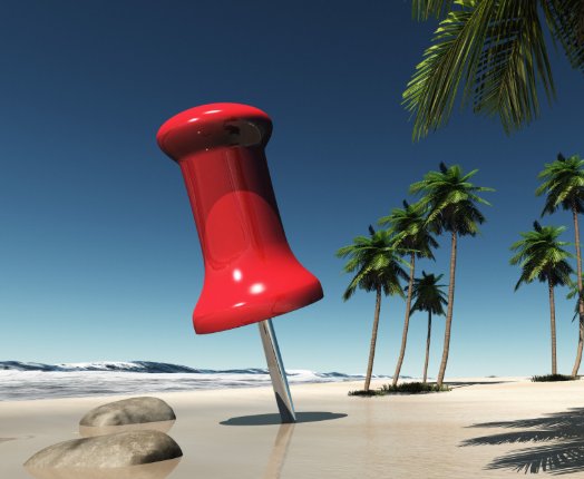 A giant, glossy red pushpin is stuck into a sandy beach beside palm trees and gentle waves under a clear blue sky.