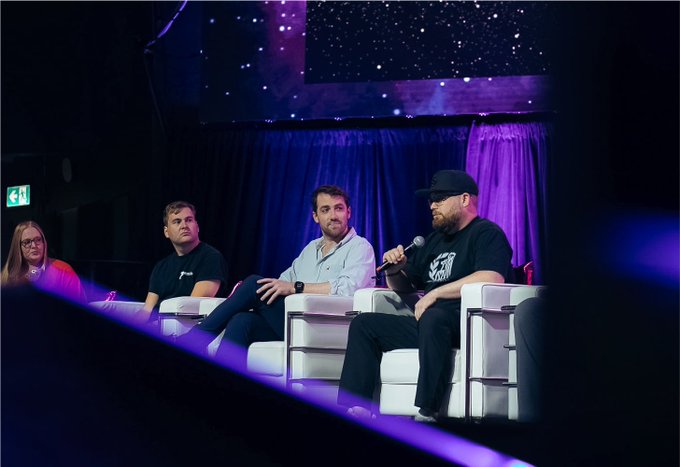 Four people seated on white chairs on a stage during a panel discussion, with a starry purple-lit backdrop and one person holding a microphone.