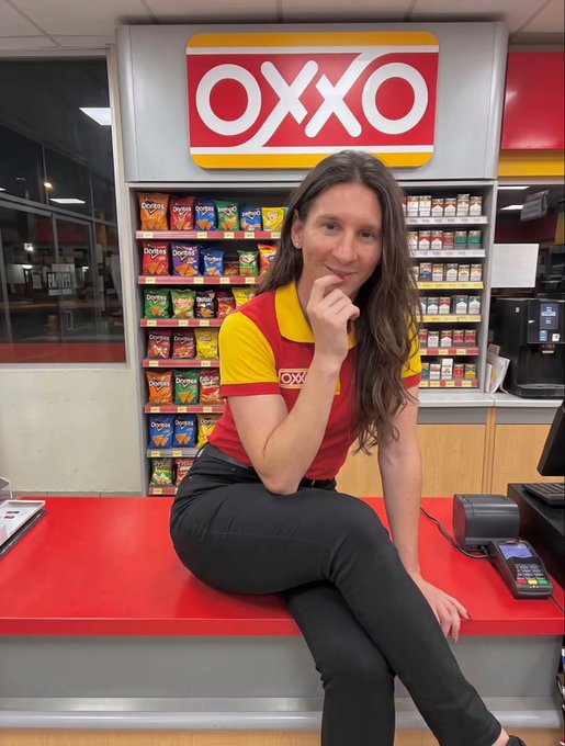 A woman in an OXXO (convenience store) uniform sits on the red counter at the checkout area, with the OXXO logo visible on the wall behind her alongside shelves of chips and beverages.