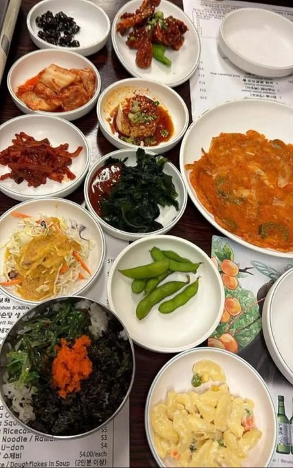 A spread of Korean dishes including kimchi, edamame, seaweed, japchae, fish soup, spicy glazed ribs, rice with vegetables, and macaroni salad arranged around a menu showing items like "U-don $4" and "Doughfricases in Soup (강된장) $4.00," fitting the caption's theme of a Korean dinner with family in Houston.

The #XRP tag in the post appears unrelated to the image content, as there are no visible cryptocurrency references, prices tickers, or financial elements shown among the food.