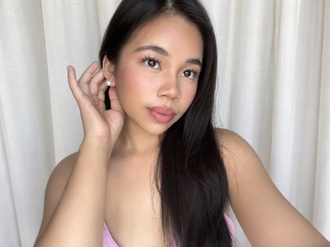 A young woman with long dark hair and natural makeup takes a mirror selfie, touching her pearl earring while wearing a pink top against a white curtain backdrop.