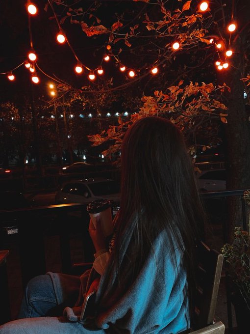 A person with long brown hair sits holding a coffee cup, viewed from behind under a canopy of glowing orange string lights at night.