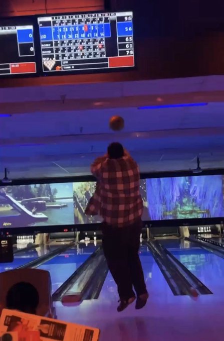 A person in a red plaid shirt bowling at a bowling alley, captured mid-throw with a ball suspended in the air, with digital scoreboards displaying frames above the lanes.