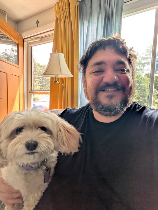 A selfie of a smiling man with a beard holding a small fluffy white and tan dog, sitting in front of a window with yellow curtains and a lamp.