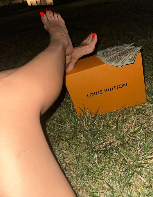 Bare legs and red-painted toes rest on an orange "LOUIS VUITTON" shopping box while a fan of cash sits on top, set in grass — a tongue-in-cheek take on a "standard" shopping haul.