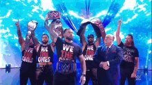 The image features a group of men standing together with their belts crossed over each other, holding up their belts as if posing for a picture. The text "We're One" can be seen on one of the shirts they are wearing. In addition to the men and their belts, there is also a man in a suit present in the image. The background appears to be a stage or a performance area, which could suggest that this group of men might be part of a wrestling team or participating in some sort of event related to wrestling.