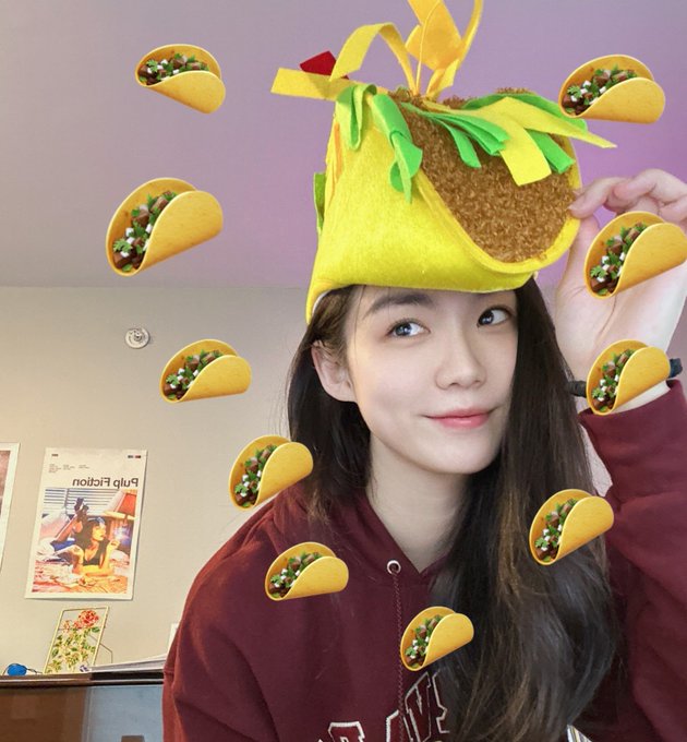 A selfie of a smiling young woman with long dark hair wearing a maroon hoodie and a creative taco-shaped hat, with digital taco stickers floating around her head; a poster in the background reads 'Blue Edition'.