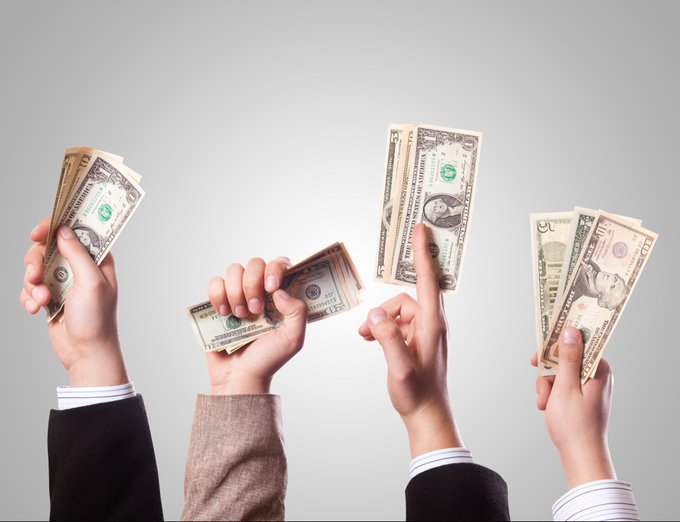 Four hands hold up stacks of US dollar bills (including $5 and $20 denominations) against a gray background.