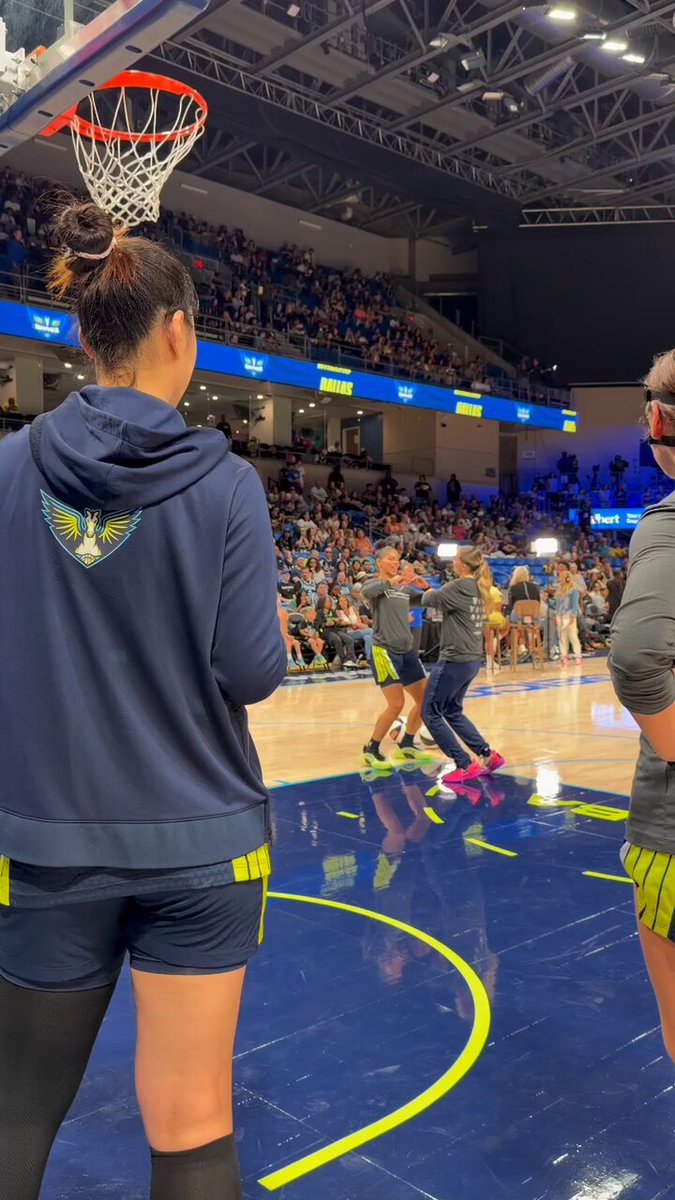 The image shows a basketball game taking place inside an indoor arena with a crowd of people watching from various positions around the court. A player wearing a blue hoodie stands near the basket, while another player can be seen walking by. The players are likely preparing for or just finished their game and are now socializing before or after the match.