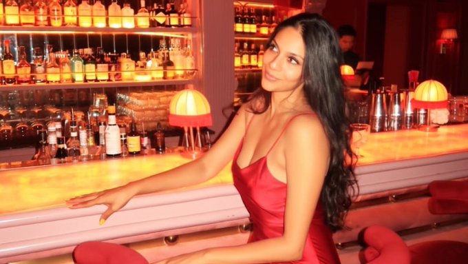 Woman with long dark hair in a red dress posing at a warmly lit bar with shelves of liquor bottles in the background.