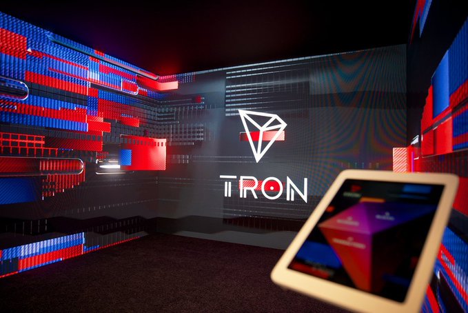 A dark, stylized promotional image for 'TRON' cryptocurrency featuring the TRON diamond logo in white against a backdrop of red and blue illuminated server-like structures, with a tablet displaying blockchain data visible in the foreground.