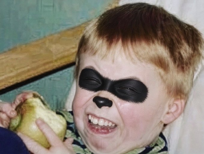 A vintage photo shows a laughing toddler with black face paint painted around their eyes and nose, holding a yellow object.