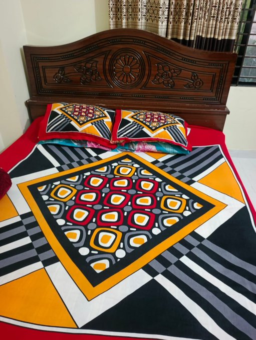 A wooden bed featuring an ornately carved headboard, dressed with a vibrant geometric-patterned bedsheet in red, black, white, yellow, and orange, along with matching pillows.
