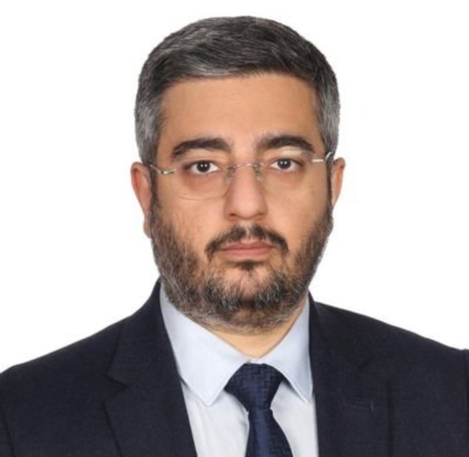 A formal portrait of a middle-aged man with a beard, glasses, and dark suit, likely representing the commenter who dismissively refers to himself in the third person as a "muşmula-suratlı" (medlar-faced) figure, humorously calling the market a "1990 model re-candidate for prosperity" while insisting Bitcoin will hold above 75k and continue rising.