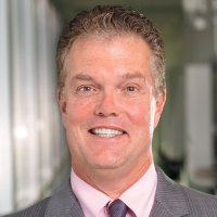 A portrait shows a smiling man with short blonde hair, wearing a gray suit, pink shirt, and dark tie.