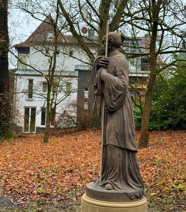 A stone statue of a robed figure holding a staff, set in an outdoor park with autumn leaves and a white building in the background.
