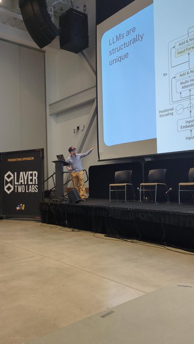 A speaker presents on stage at a conference, with a slide titled "LLMs are structurally unique" displayed on a large screen beside them. A banner for "Layer Two Labs" is visible on the left, matching the conference setting referenced in the caption as @tabconf, where the presenter is discussing LLMs and about to introduce L402s.