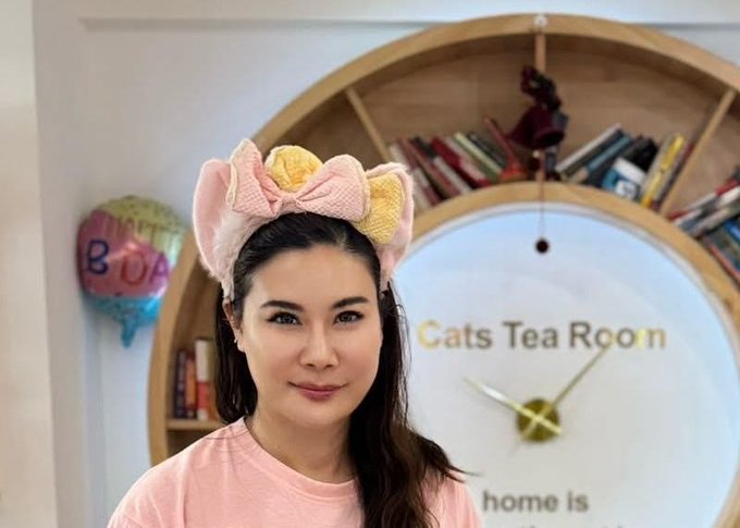 A woman with dark hair wears a pink and yellow cat-ear headband, smiling in front of a wooden shelf featuring a clock labeled "Cats Tea Room" and a "Happy Birthday" balloon.
