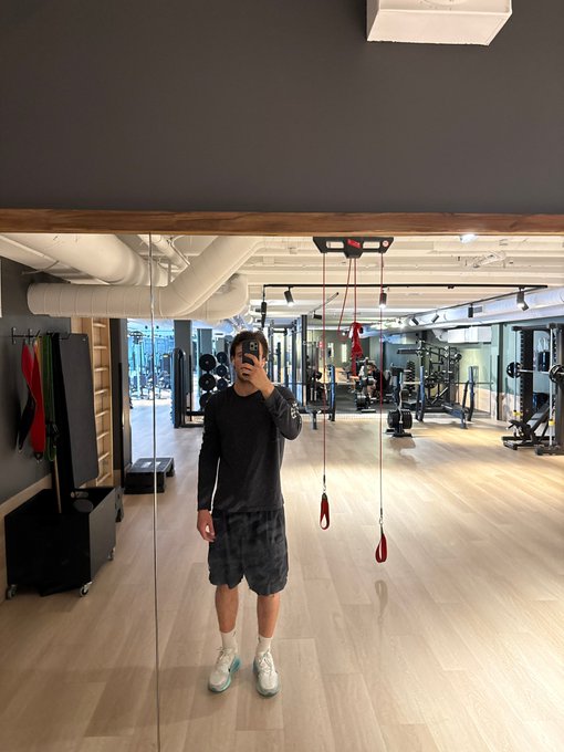 A person takes a mirror selfie in a gym, wearing a dark long-sleeve shirt, patterned shorts, white socks, and light-colored sneakers, with various workout equipment visible in the background.