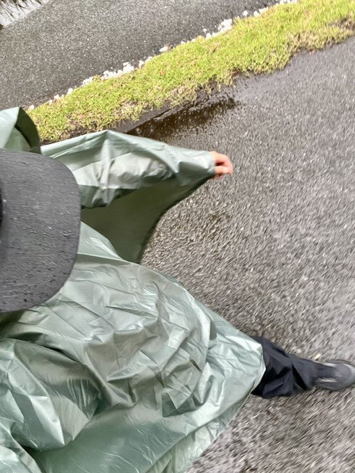 The image shows a person wearing a gray raincoat, walking down a street with their hand outstretched. The photo appears to have been taken during a rainy day, as evidenced by the wet ground and the person's attire. The caption of the image reads "ポンチョ無駄な面積多いな。デザインかな". This phrase is likely an inside joke or reference to a specific event that occurred during the rainy day captured in the photo.