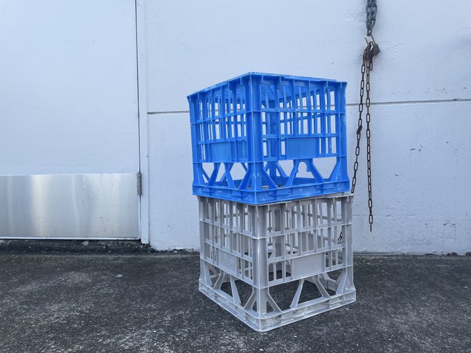 A blue plastic crate stacked on top of a gray plastic crate, sitting on concrete next to a white wall with a metal chain hanging down.