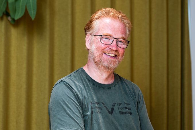 An older man with reddish-blonde hair and glasses smiles warmly while wearing a green graphic t-shirt, photographed against olive green curtains.