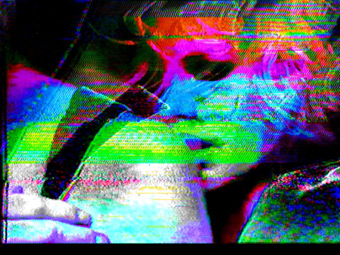A heavily distorted, glitchy digital image featuring colorful artifacts in red, blue, green, and purple across an unrecognizable scene.