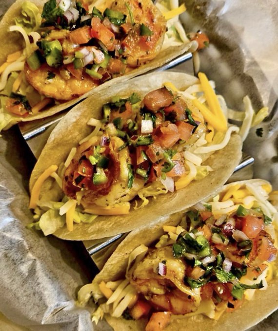 Three loaded shrimp tacos sit on parchment paper over a wire rack, generously topped with shredded cheese, diced tomatoes, onions, and cilantro.