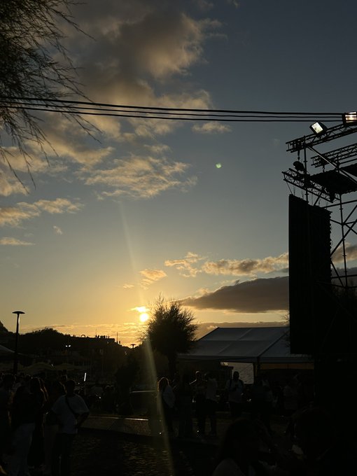 A crowd gathers at sunset beneath scattered clouds at what appears to be an outdoor event with a large sound system and lighting rig silhouetted against the golden sky, tying into the post's message about a profitable day of transactions or deals.