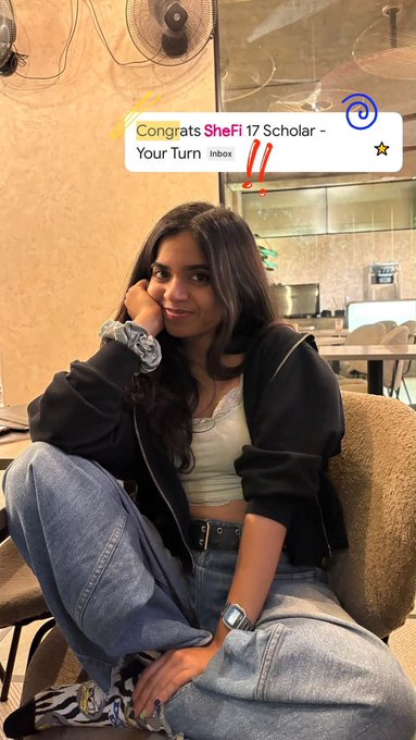 A young woman with long dark hair, wearing a black jacket and jeans, sits in a chair with a Snapchat story notification reading "Congrats SheFi 17 Scholar - Your Turn" displayed above her.