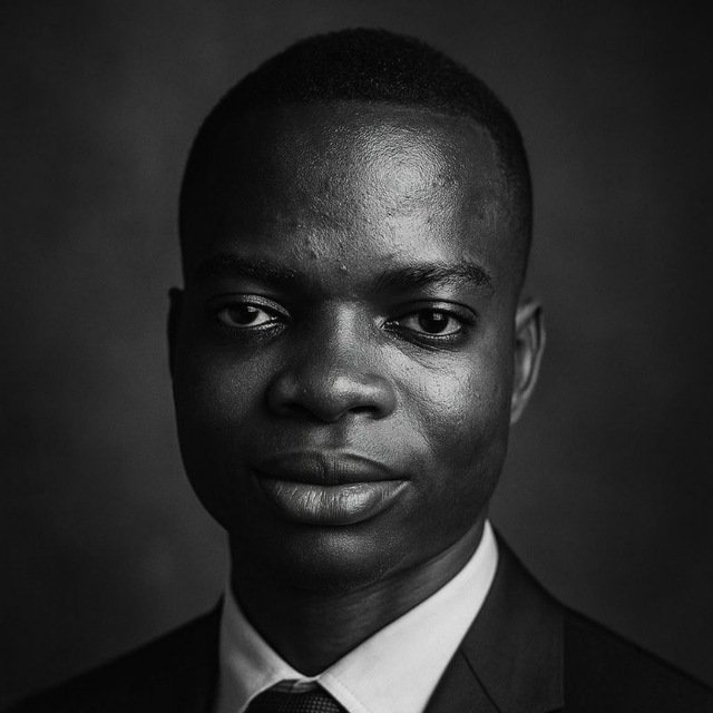 A black-and-white studio portrait of a man in a suit and tie, looking directly at the camera with a neutral expression.