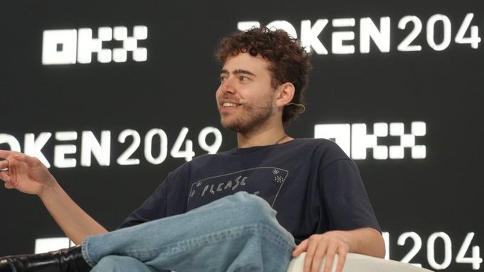 A young man with curly brown hair and a beard, wearing a dark t-shirt, speaking on stage at the OKX TOKEN 2049 conference.