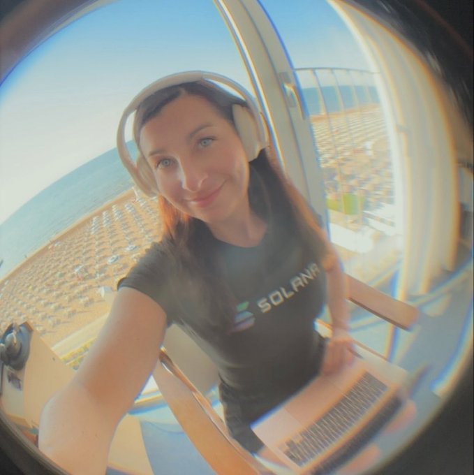 A fisheye lens selfie of a smiling woman wearing headphones and a black 'SOLANA' t-shirt, sitting on a balcony overlooking a beach and city skyline.