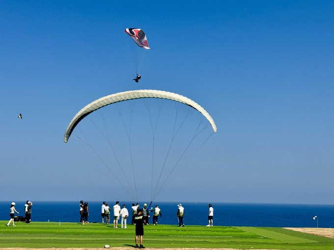 A paraglider soars above a coastal cliff where spectators watch against a clear blue sky and ocean backdrop, paired with a promotional crypto price prediction post tagged with hashtags #BTC, #SOL, #TAO, #SUI, and #XRP alongside a sponsorship from @binancezh. The image's soaring flight and open horizon visually echo the aspirational "moonshot" crypto targets advertised.