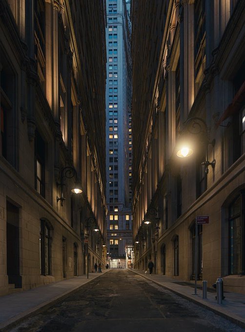 A narrow urban alleyway flanked by tall, illuminated buildings leading toward a distant skyscraper at dusk.